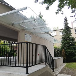 Dariusz Stepa METAL-HOUSE - Wejście do budynku z balustradą schodową i szklanym zadaszeniem wspartym na metalowych elementach mocujących, widok z zewnątrz.