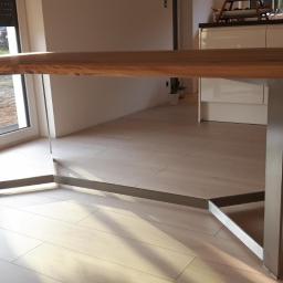 Dariusz Stepa METAL-HOUSE - Stół z blatem z jasnego drewna i nogami z nierdzewnej stali o geometrycznym kształcie, widok z boku na tle jasnej podłogi i okna z widokiem na zewnątrz.