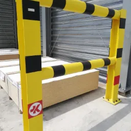 Żółta metalowa bariera ochronna z czarnymi pasami i logo producenta, zabezpieczająca stos płyt drewnianych w hali magazynowej.