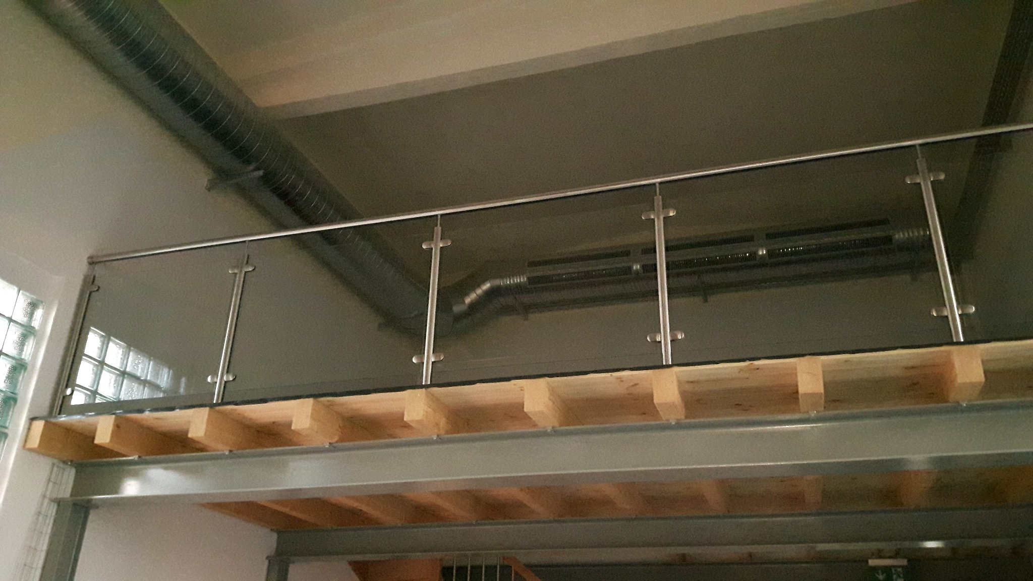 Szklana balustrada z nierdzewnymi słupkami na antresoli w industrialnym wnętrzu, widoczne drewniane belki konstrukcyjne i metalowe rury wentylacyjne pod sufitem.