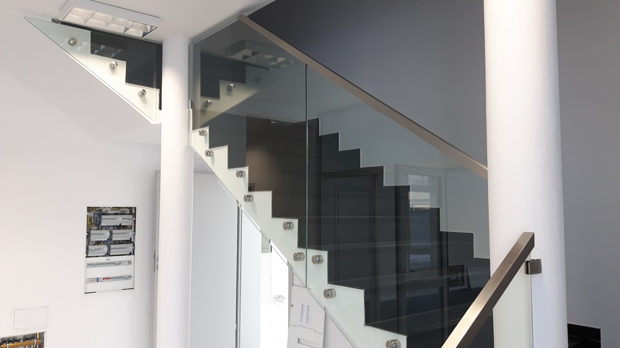 Nowoczesne schody wewnętrzne z czarnymi stopniami i szklaną balustradą mocowaną na metalowych uchwytach, widoczne z boku, z kolumną nośną i skrzynką elektryczną w tle.