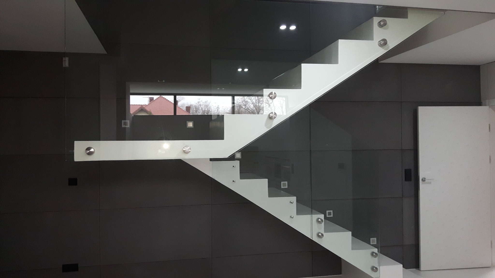 Nowoczesne schody metalowe z białymi stopniami i szklaną balustradą, mocowane na metalowych śrubach, zrealizowane w Żarach, widoczne na tle ciemnoszarej ściany i białych drzwi.