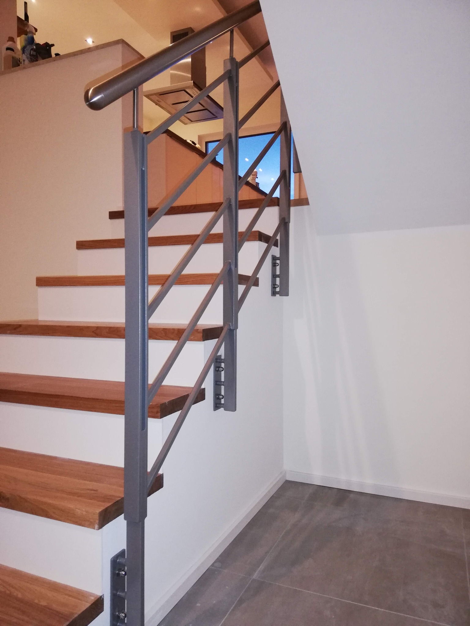 Metalowa balustrada schodowa w kolorze szarym, z drewnianymi stopniami, zamocowana do białej ściany, widok z dołu na górę schodów.