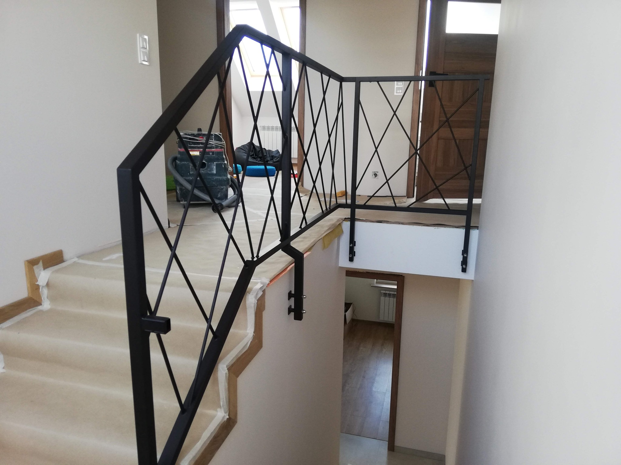 Nowoczesna, czarna balustrada metalowa z motywem krzyżowym, zamontowana na schodach w trakcie remontu, zabezpieczonych folią malarską, widoczny odkurzacz przemysłowy w tle.