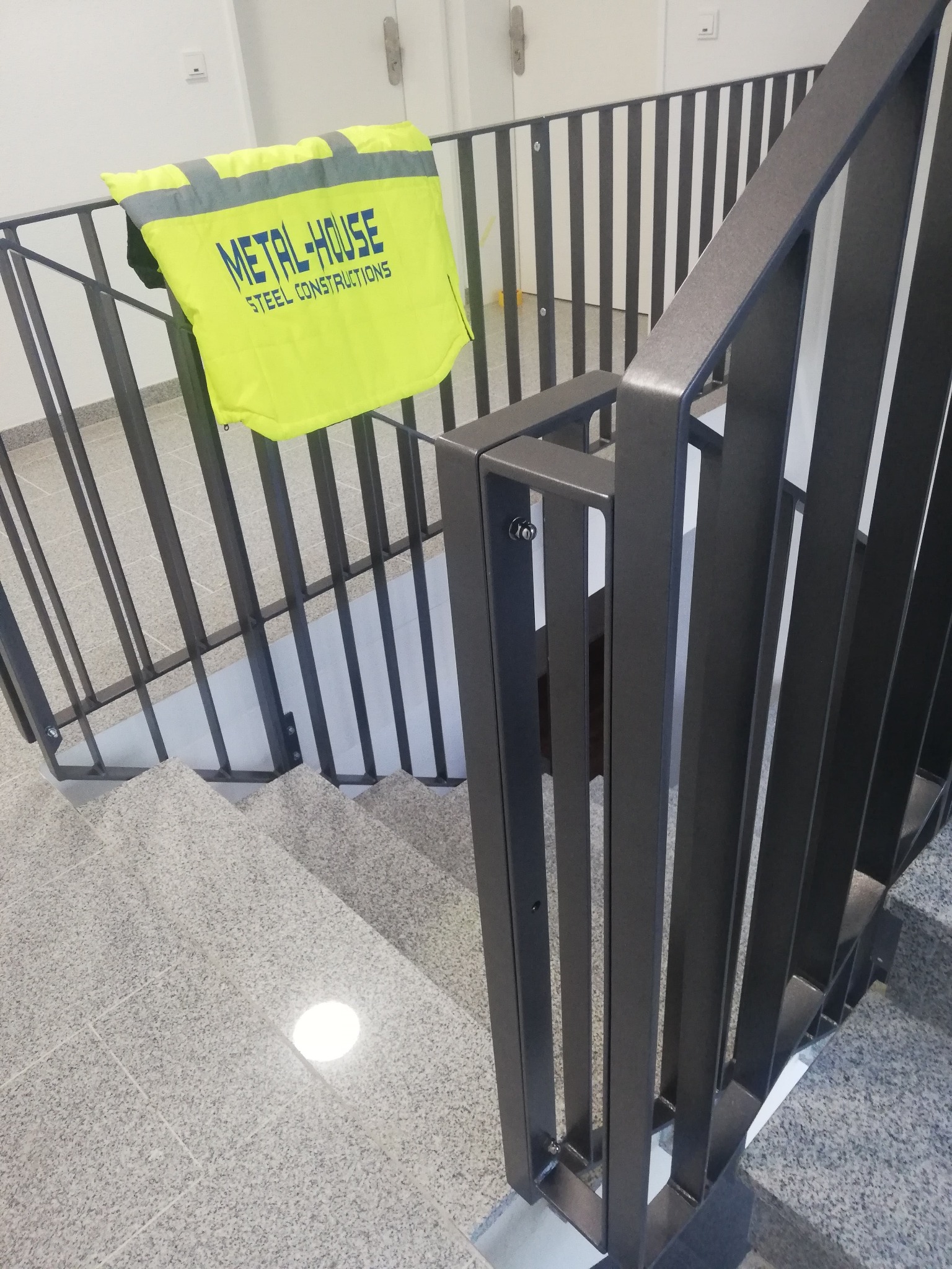 Nowoczesna, metalowa balustrada schodowa wewnątrz budynku, z żółtą kamizelką odblaskową firmy Metal-House zawieszoną na poręczy.