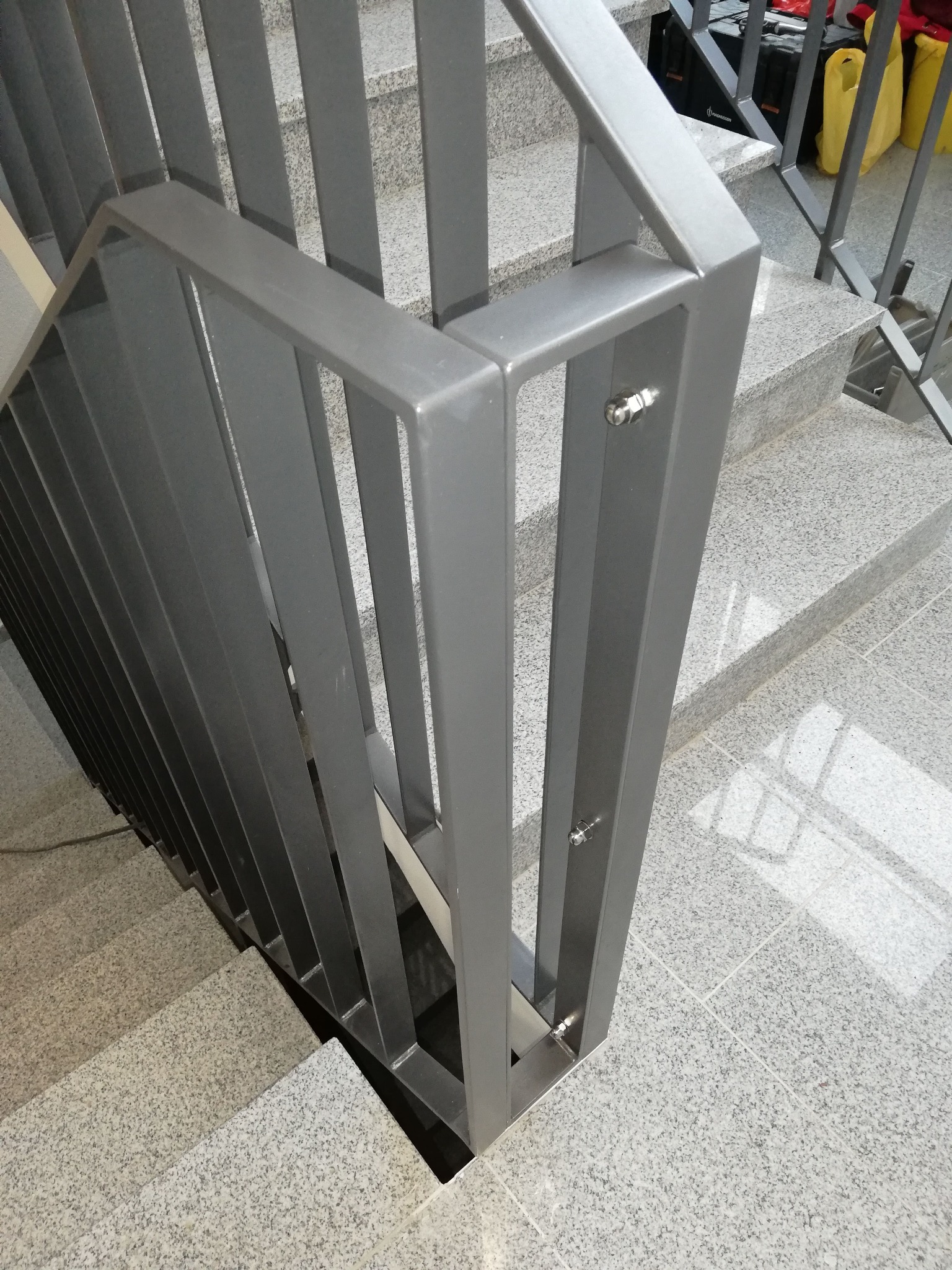 Szare schody z granitowych płytek z nowoczesną, metalową balustradą o prostych, pionowych szczeblach, mocowaną na śruby.