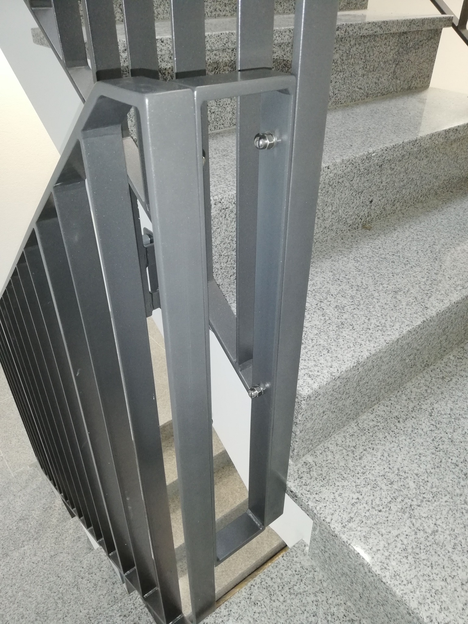 Szare metalowe schody z minimalistyczną balustradą z pionowych płaskowników, mocowaną śrubami do stopni z szarego lastryko.