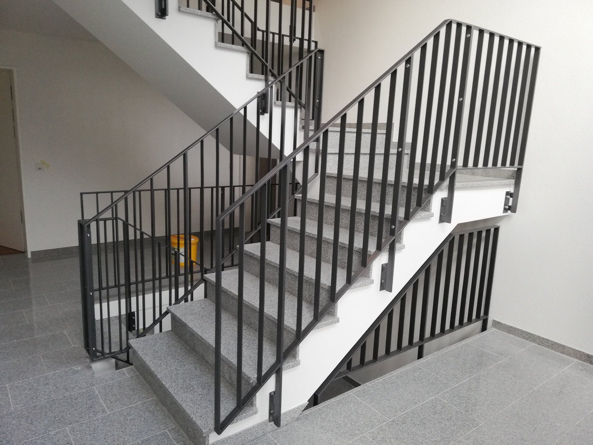 Wewnętrzne schody z szarymi, kamiennymi stopniami i czarną, metalową balustradą z pionowych prętów, widoczne z dołu, z żółtym wiadrem w tle.