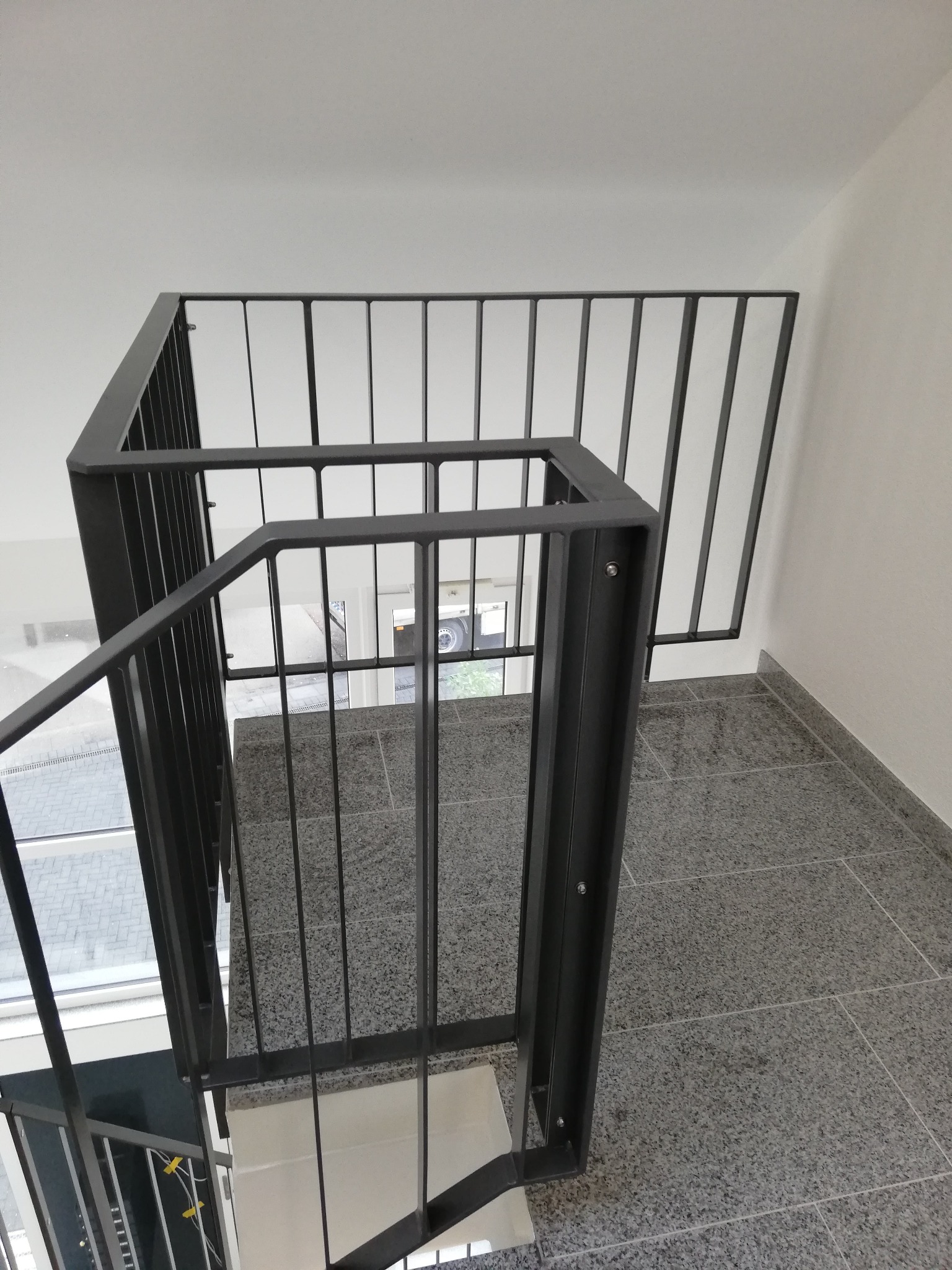 Widok z góry na nowoczesną, metalową balustradę schodową o minimalistycznym designie, z widocznymi śrubami mocującymi, na tle szarej, kamiennej podłogi i białych ścian.