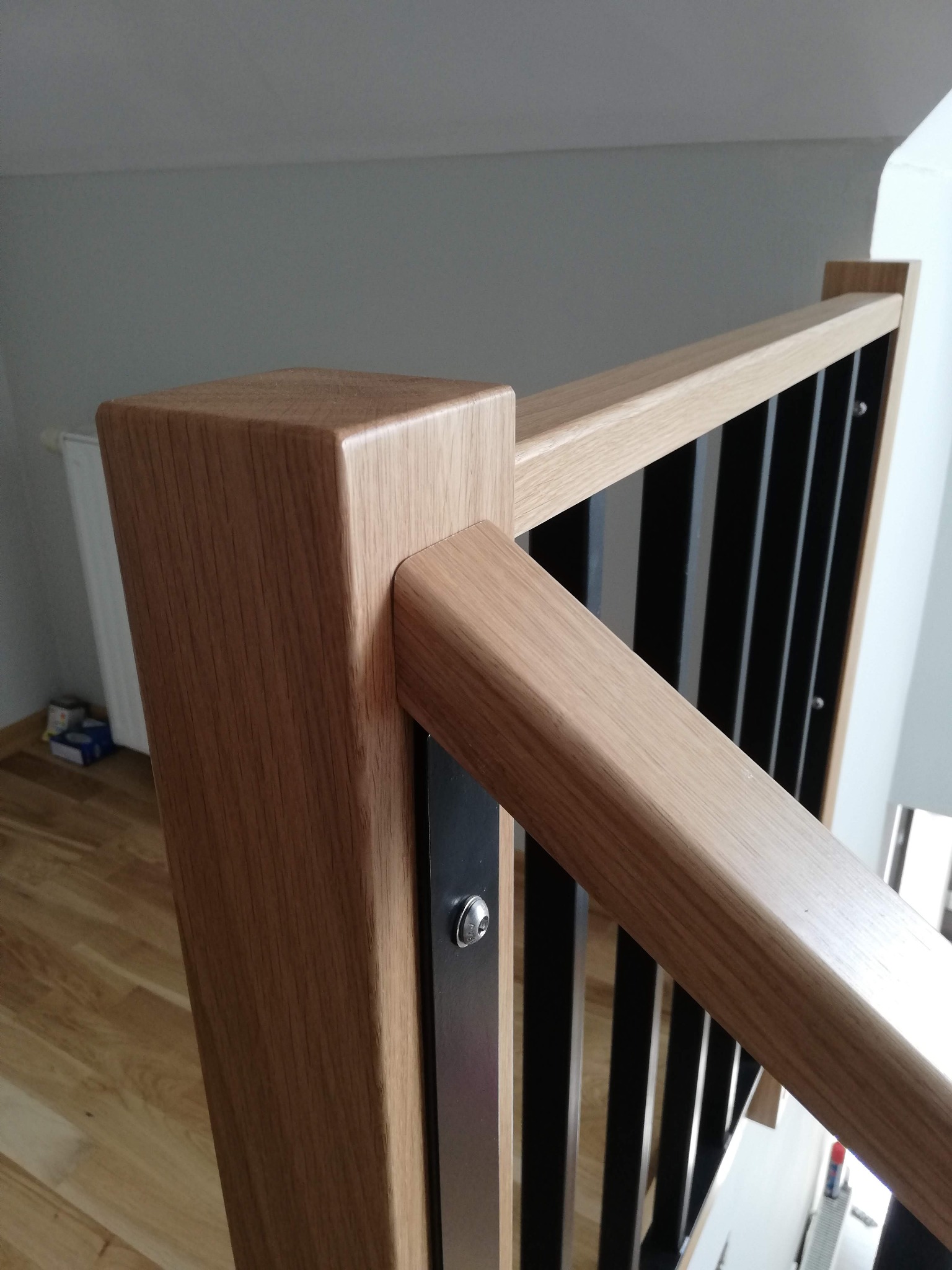 Fragment balustrady schodowej z drewnianymi elementami w odcieniach beżu i czarnymi, pionowymi słupkami, mocowanymi śrubami, widok z góry na podłogę.
