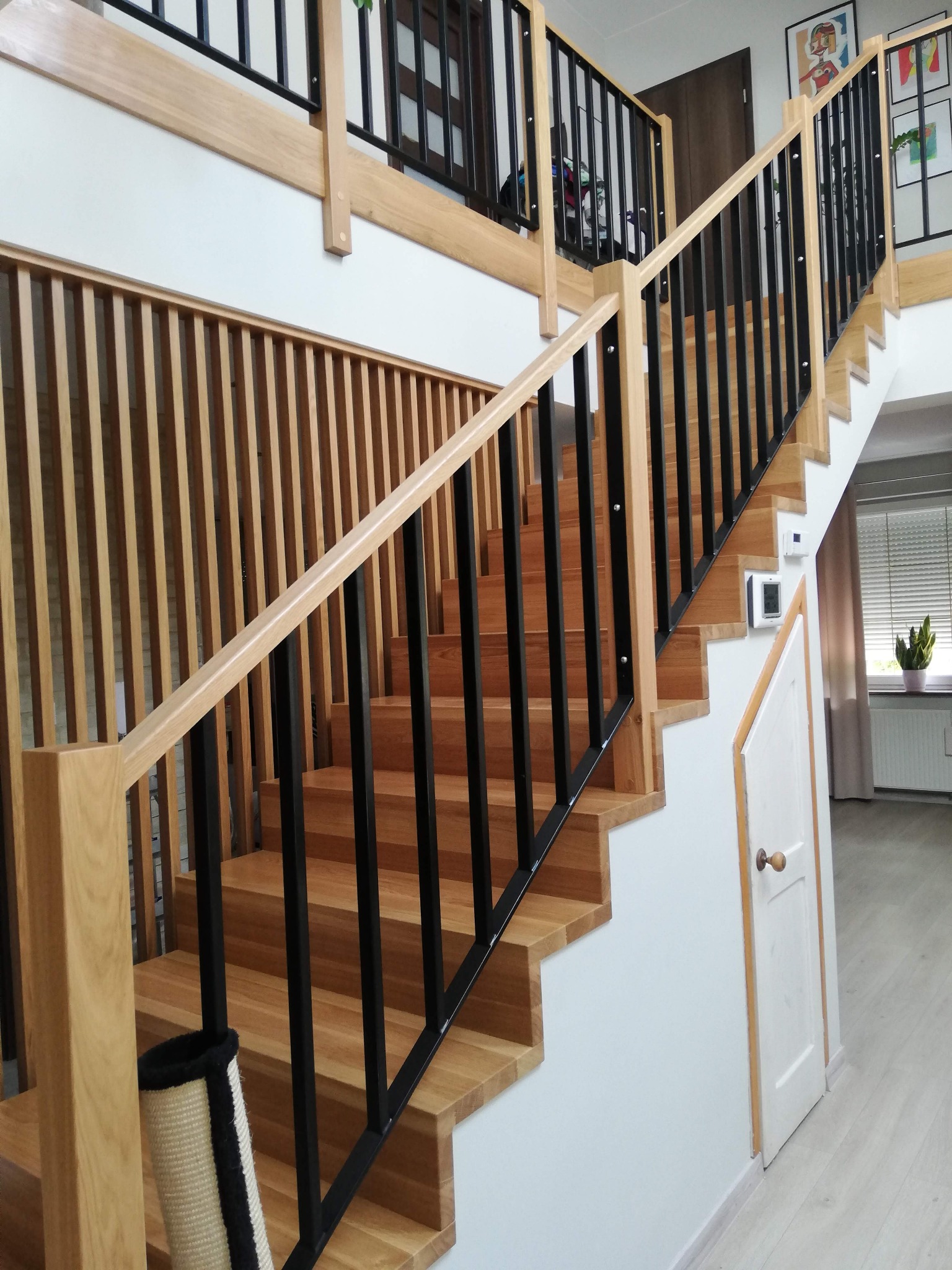 Drewniane schody z czarnymi metalowymi balustradami w nowoczesnym wnętrzu, widoczne elementy wykończenia z jasnego drewna i białe ściany.