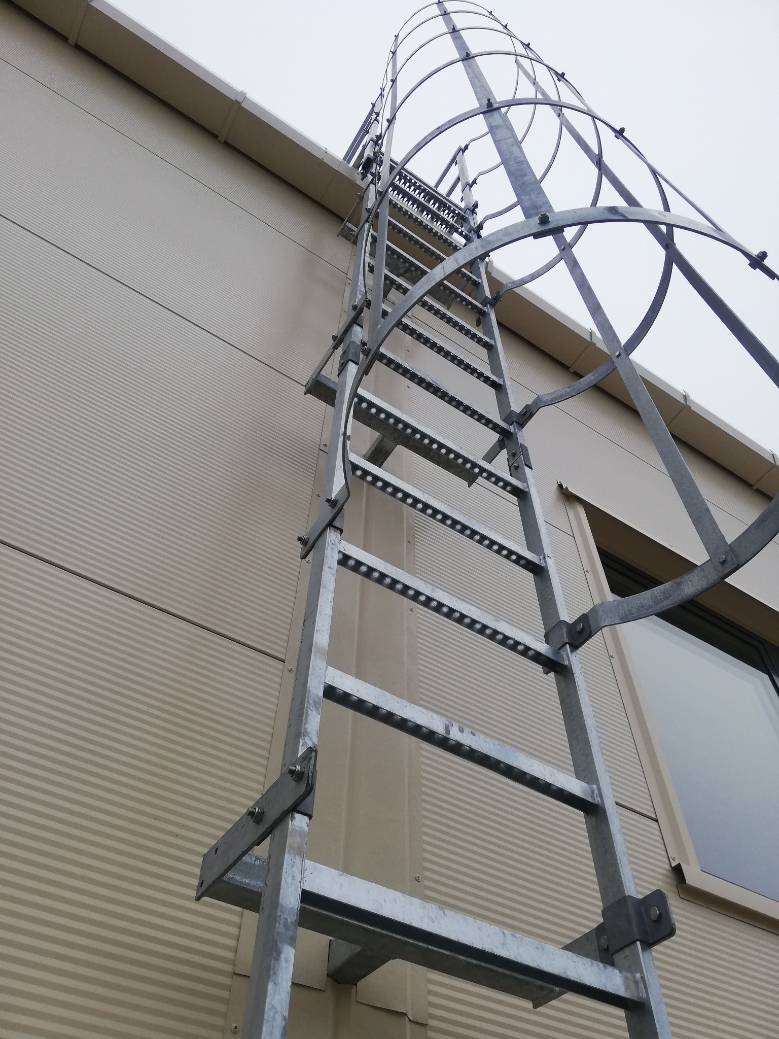 Zewnętrzna metalowa drabina ewakuacyjna z koszem ochronnym przymocowana do ściany budynku z blachy trapezowej, widoczna z dołu pod kątem.