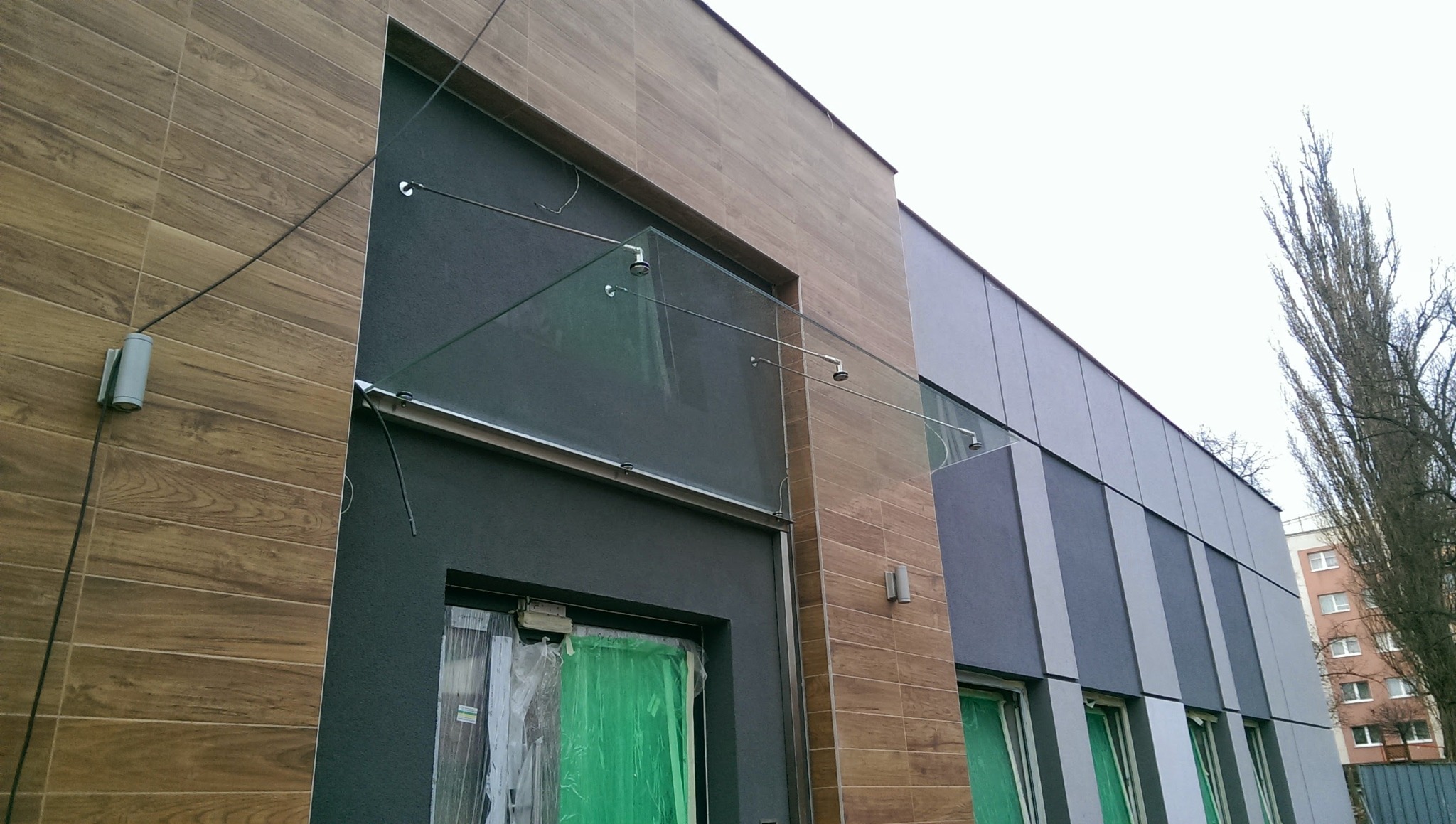 Szklany daszek nad wejściem do budynku z drewnopodobną elewacją, podparty metalowymi wspornikami, widoczne okna zabezpieczone folią ochronną.