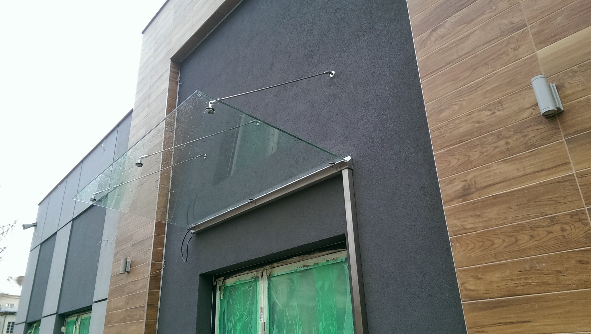 Szklany daszek nad wejściem do budynku, wsparty na metalowych wspornikach, z widocznymi kroplami deszczu na szkle i zabezpieczonymi folią oknami poniżej.