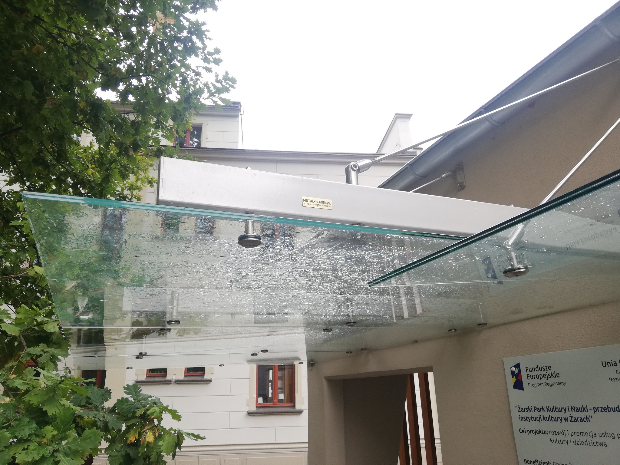 Szklany daszek nad wejściem do budynku z widocznymi kroplami deszczu, wsparty na metalowej konstrukcji mocowanej do ściany, z zielenią drzew w tle.
