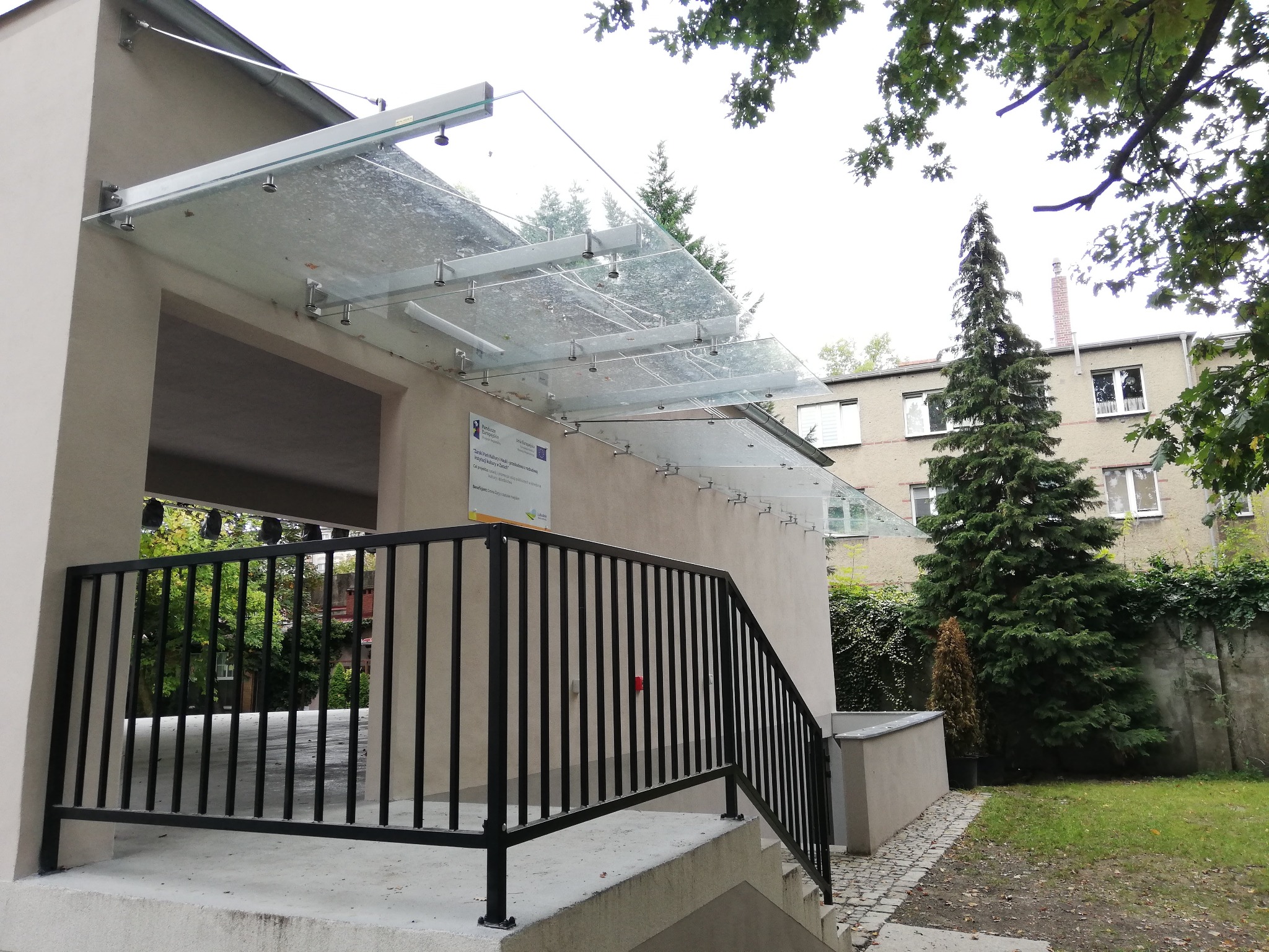 Wejście do budynku z balustradą schodową i szklanym zadaszeniem wspartym na metalowych elementach mocujących, widok z zewnątrz.