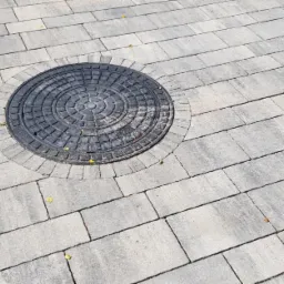 Szara kostka brukowa ułożona wokół studzienki kanalizacyjnej. Widoczne niewielkie, żółte liście na powierzchni.