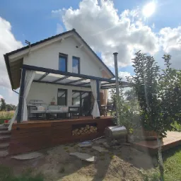 Nowoczesny taras z drewna z pergolą i zasłonami, idealny do relaksu na świeżym powietrzu. Widok na zadbany ogród i błękitne niebo z chmurami.