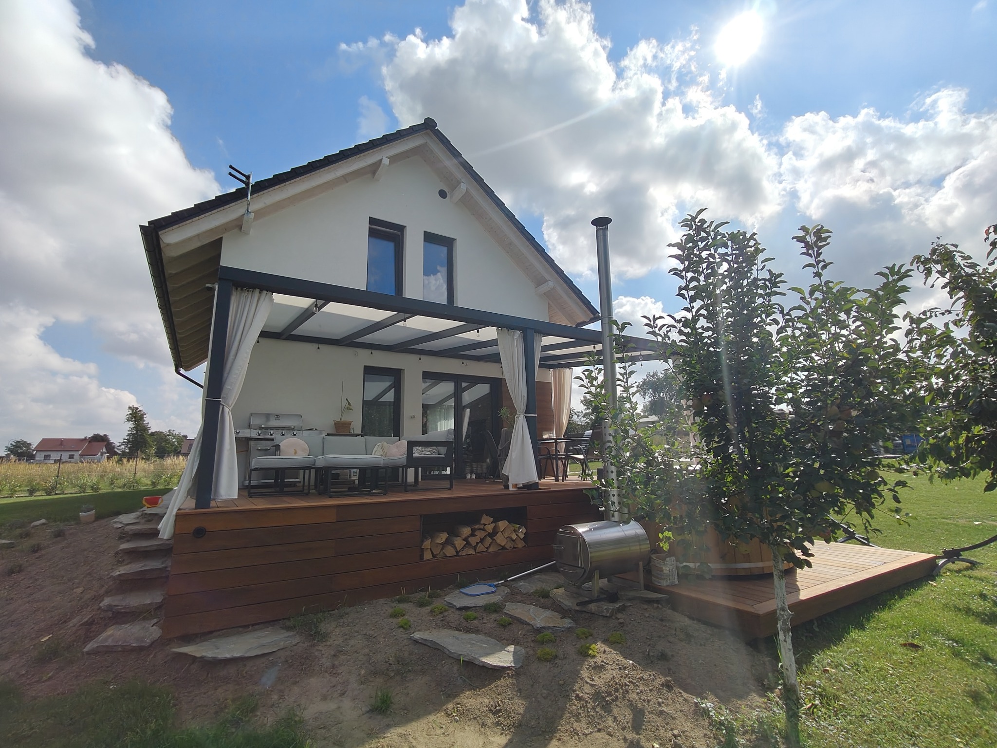 Nowoczesny taras z drewna z pergolą i zasłonami, idealny do relaksu na świeżym powietrzu. Widok na zadbany ogród i błękitne niebo z chmurami.