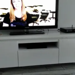 Biała, lakierowana szafka RTV z metalowymi nóżkami, na blacie telewizor Samsung i dekoder, podłoga wyłożona płytkami imitującymi drewno.