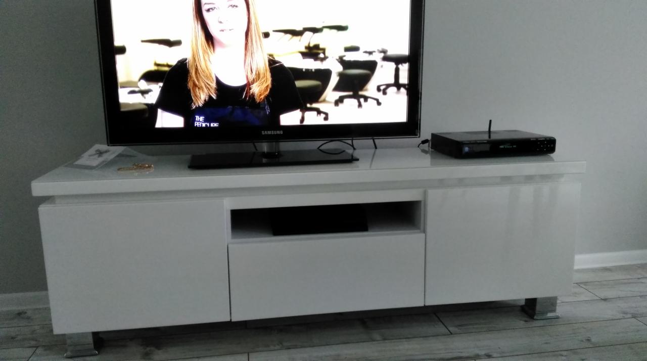 Biała, lakierowana szafka RTV z metalowymi nóżkami, na blacie telewizor Samsung i dekoder, podłoga wyłożona płytkami imitującymi drewno.