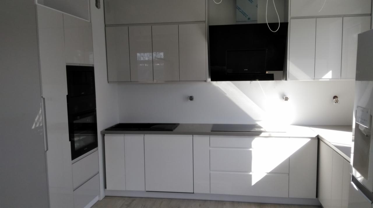 Nowoczesna, biała kuchnia z błyszczącymi frontami szafek, czarnym panelem nad blatem i wbudowanym sprzętem AGD, oświetlona naturalnym światłem wpadającym przez okno.