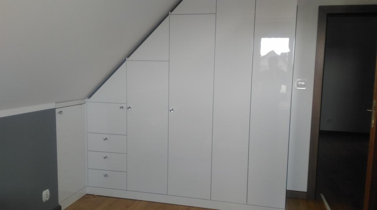 Biała zabudowa meblowa pod skosem dachu z szafkami i szufladami, widoczny fragment podłogi i drzwi do innego pomieszczenia.