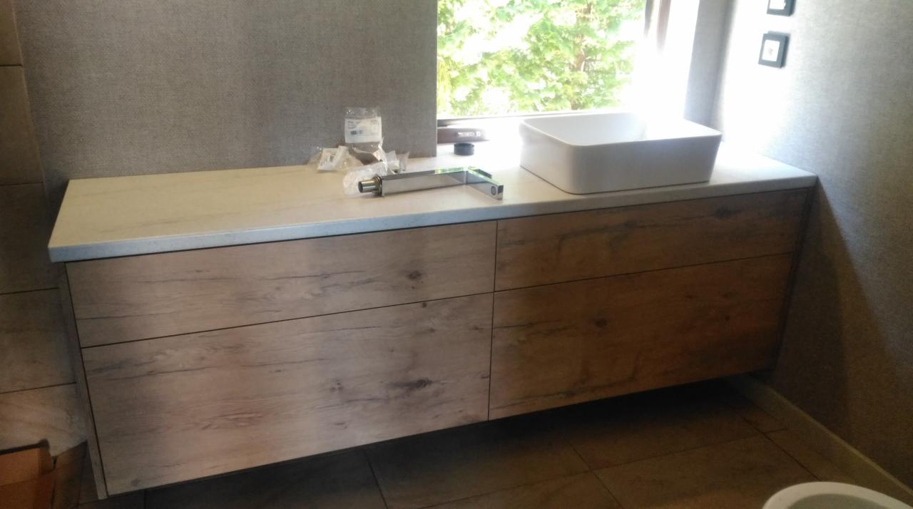 Łazienkowa szafka pod umywalkę z drewnopodobnym frontem, biały blat i umywalka nablatowa, bateria w trakcie montażu, widoczne elementy instalacyjne.