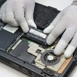 Otwarty laptop z widocznym wnętrzem, ręce w białych rękawiczkach instalują moduł pamięci RAM, wentylator chłodzenia procesora widoczny w tle.