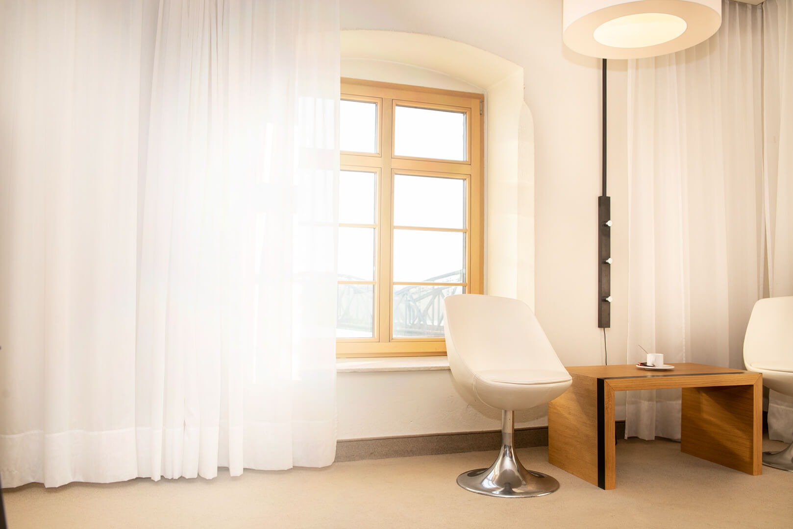 Jasne wnętrze z okrągłym oknem, krzesłem na chromowanej nodze, drewnianym stolikiem z filiżanką i lampą podłogową z okrągłym kloszem, zasłonięte firankami.