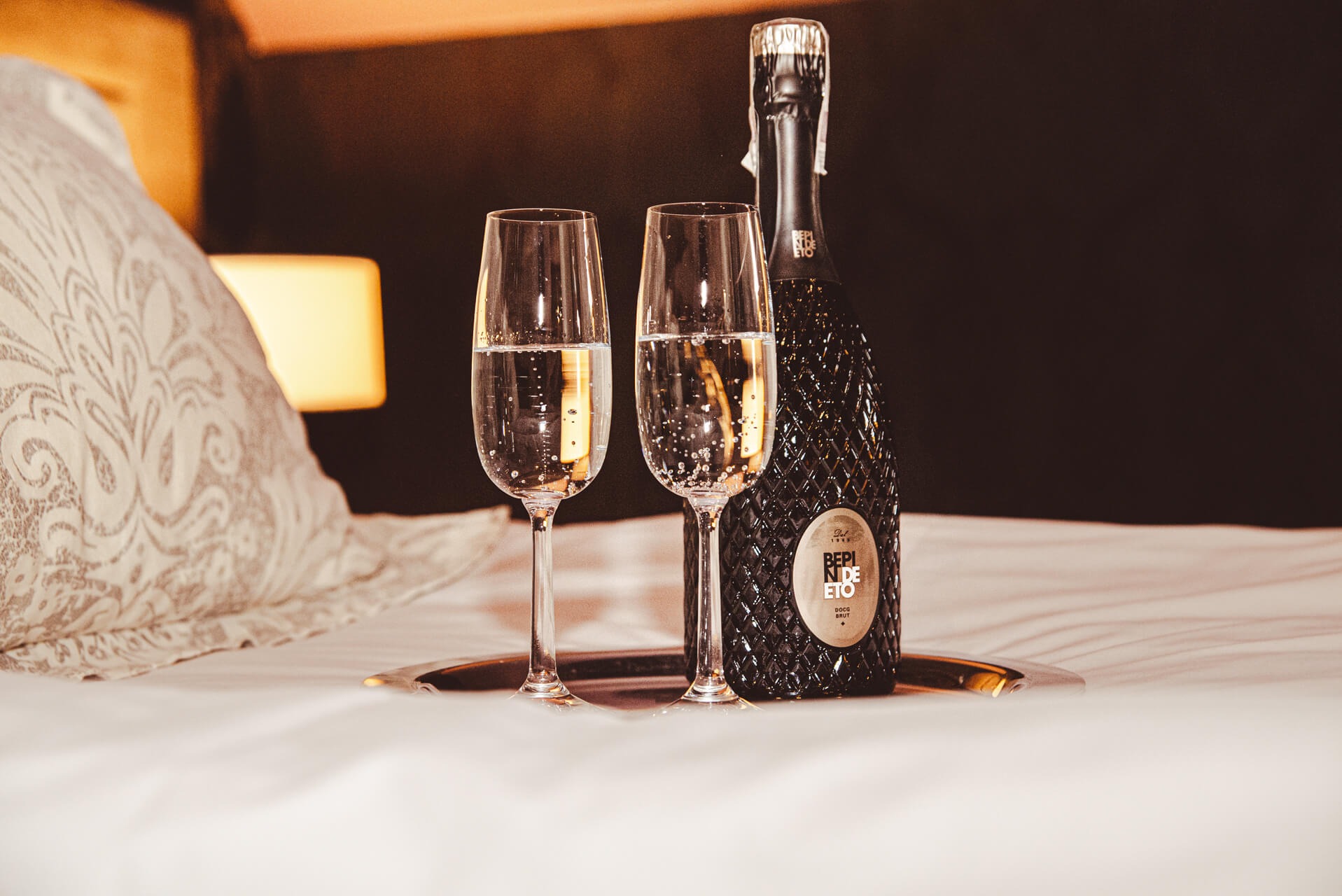 Dwa kieliszki szampana i butelka wina musującego na białej pościeli z wzorem, miękkie światło lampki nocnej w tle.