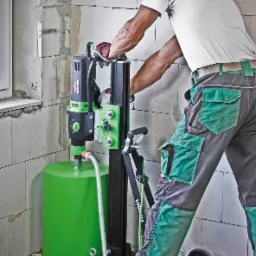 Pracownik w słuchawkach ochronnych obsługuje wiertnicę rdzeniową marki Eibenstock, wiercąc otwór w ścianie z betonu komórkowego. Widoczne ślady po wierceniu na ścianie i podłodze.