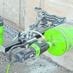 Wiertnica diamentowa Eibenstock zamocowana do ściany betonowej, wiercąca otwór. Widoczny zbiornik na wodę i system mocowania z szyną. Ślady pracy na wiertle.