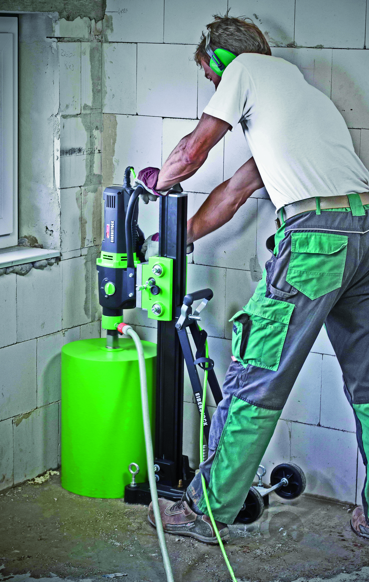 Pracownik w słuchawkach ochronnych obsługuje wiertnicę rdzeniową marki Eibenstock, wiercąc otwór w ścianie z betonu komórkowego. Widoczne ślady po wierceniu na ścianie i podłodze.