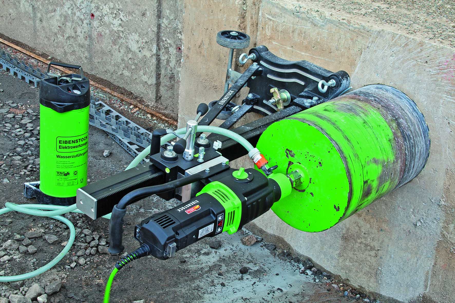 Wiertnica diamentowa Eibenstock zamocowana do ściany betonowej, wiercąca otwór. Widoczny zbiornik na wodę i system mocowania z szyną. Ślady pracy na wiertle.