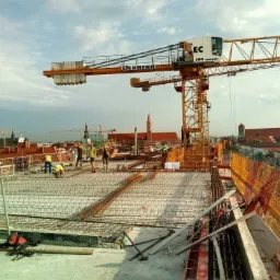 Zbrojenie stropu na budowie z widocznym dźwigiem Liebherr na tle miejskiej zabudowy z kościołami i kominami, pracownicy w żółtych kamizelkach w trakcie montażu siatki zbrojeniowej.