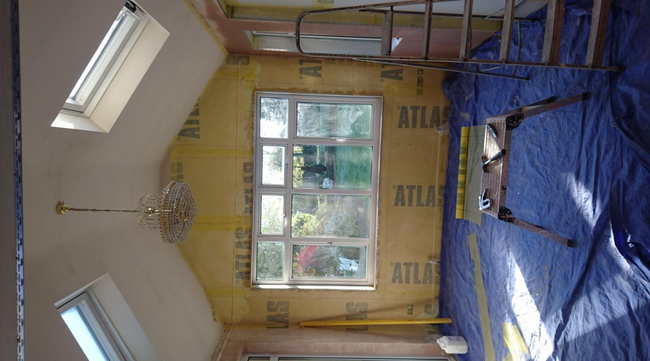 Pokój w trakcie remontu: zabezpieczone ściany płytami, okna dachowe i standardowe, wiszący żyrandol, rozłożona drabina, podłoga zabezpieczona folią.