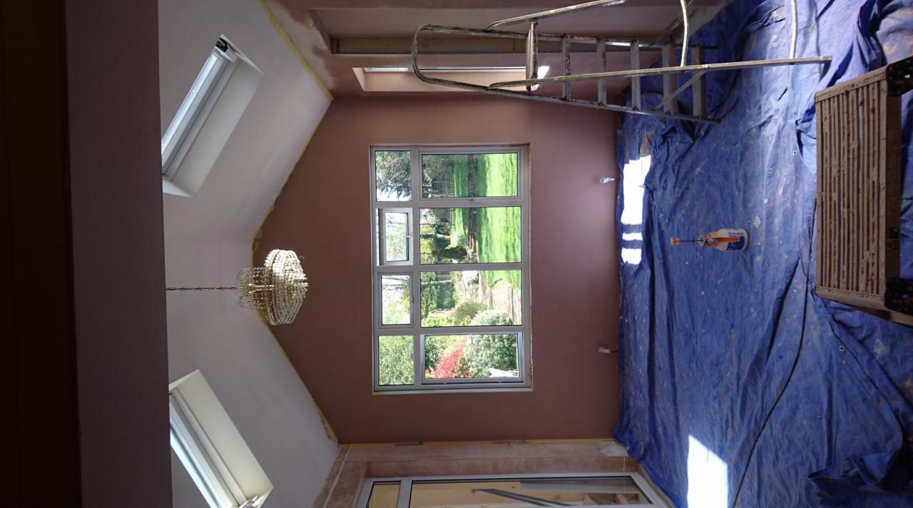 Pokój w trakcie prac wykończeniowych: ściany przygotowane pod gładź, okna dachowe i duże okno z widokiem na ogród, zabezpieczona podłoga, drabina oparta o ścianę.