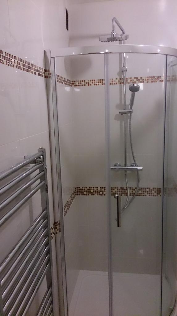 Narożna kabina prysznicowa z deszczownicą i panelem natryskowym, obok grzejnik drabinkowy, białe płytki z dekoracyjnym pasem brązowej mozaiki.