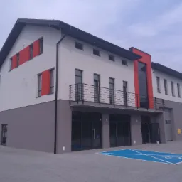 Dwukondygnacyjny budynek biurowy z elementami czerwieni na elewacji, widok z zewnątrz na szarym brukowanym placu z oznaczonym miejscem parkingowym dla niepełnosprawnych.