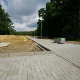 Świeżo ułożona, szara kostka brukowa w jodełkę tworząca chodnik i ścieżkę, z paletą kostki w tle, obok teren przygotowany pod dalsze prace ziemne.