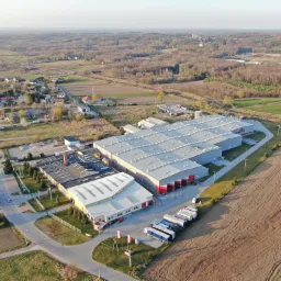 Widok z lotu ptaka na kompleks magazynowy z rampami załadunkowymi i zaparkowanymi ciężarówkami, otoczony polami i zabudową mieszkalną.