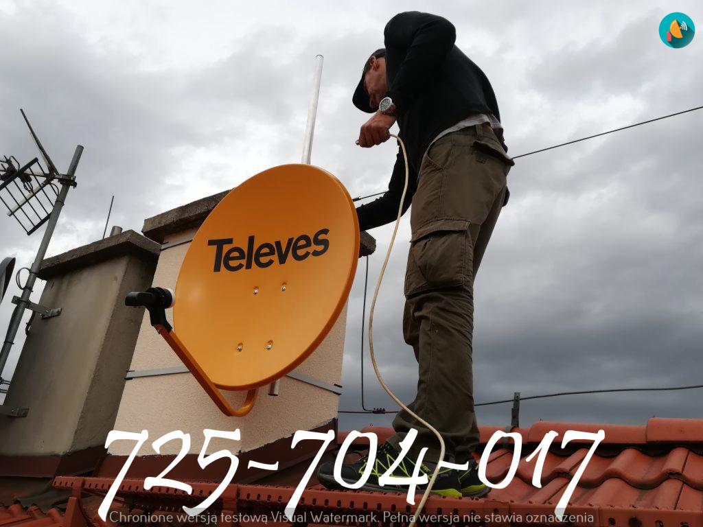 Instalator na dachu montuje pomarańczową antenę satelitarną Televes, widok z dołu na pochmurne niebo.