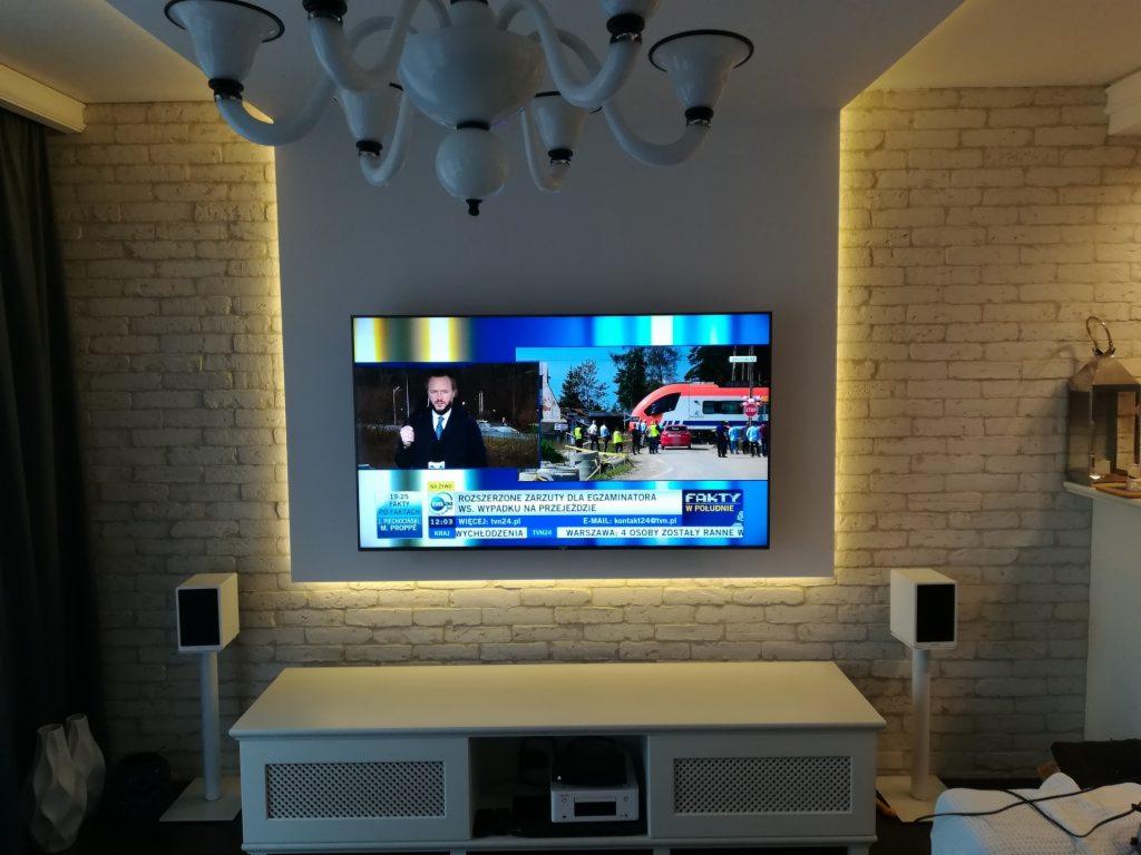 Telewizor zamontowany na ścianie z cegły, podświetlony ledami, z białą szafką RTV i głośnikami po bokach, pod dekoracyjnym żyrandolem.