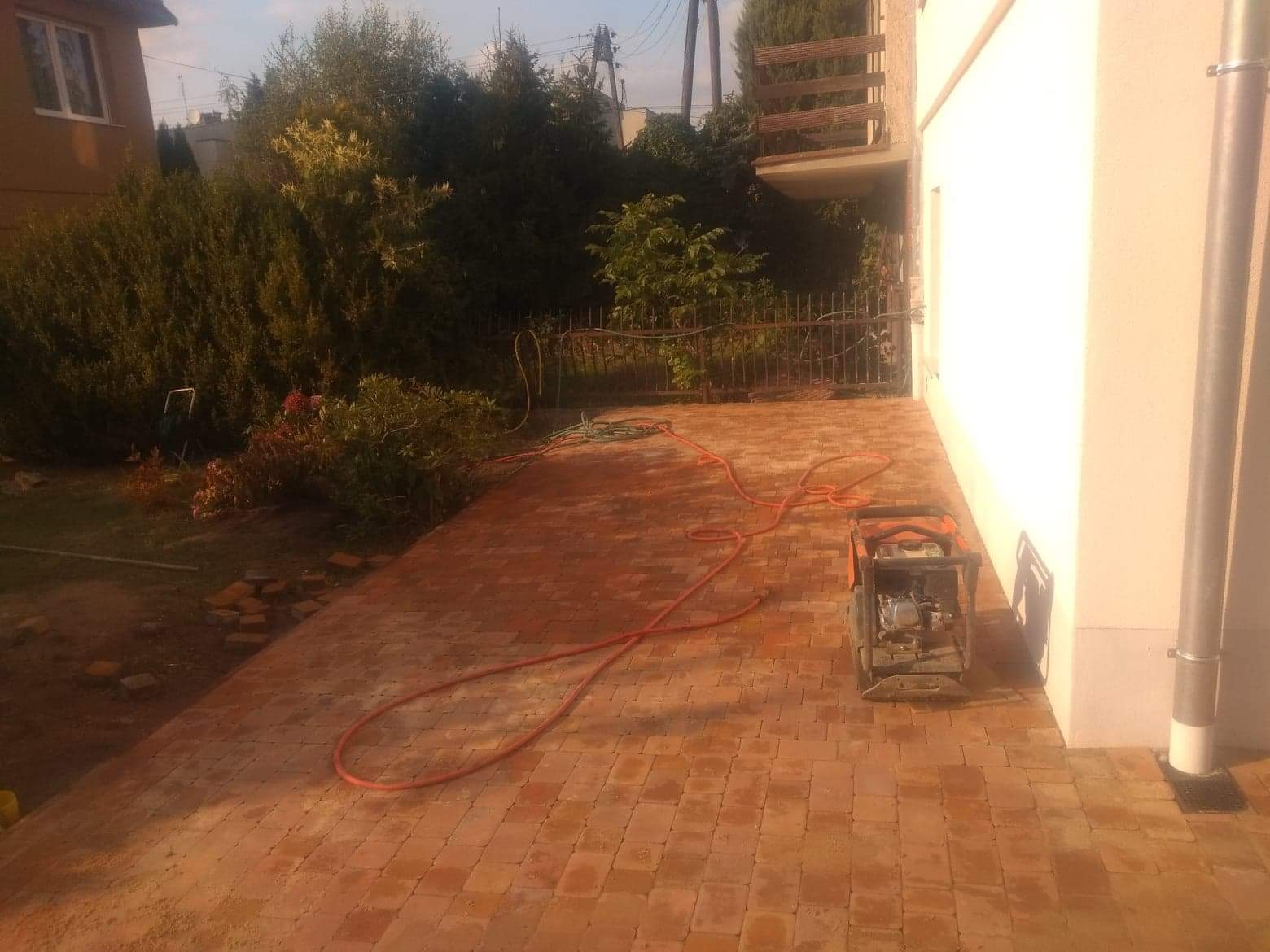 Nowo ułożony taras z ceglanej kostki, widoczny wąż ogrodowy i zagęszczarka wibracyjna, dom z białym tynkiem w tle.