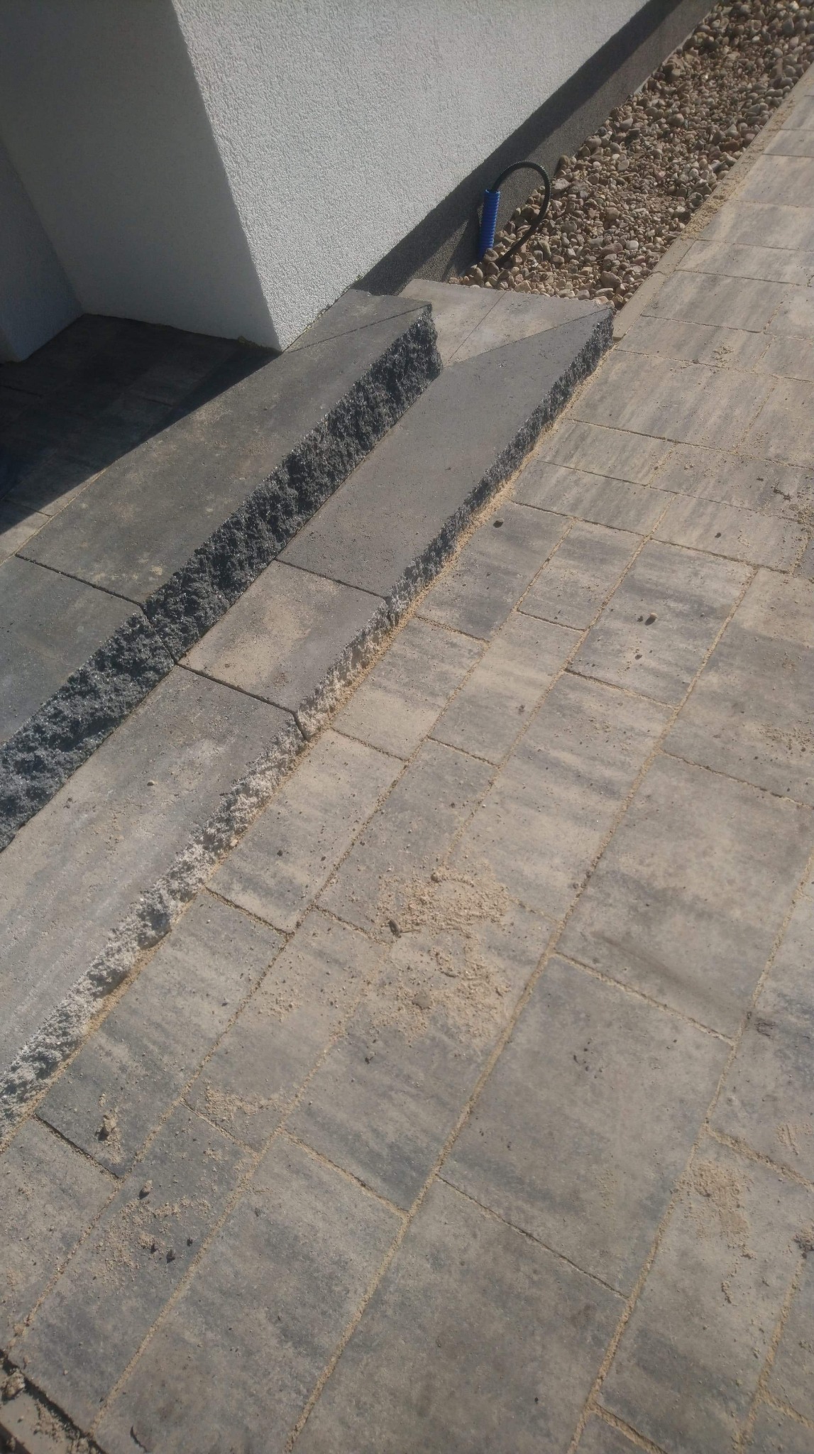 Dwa stopnie schodów z kostki granitowej prowadzące do białego budynku, obok widoczny drenaż z kamyków i fragment chodnika z kostki brukowej. Widoczne ślady piasku na kostce.