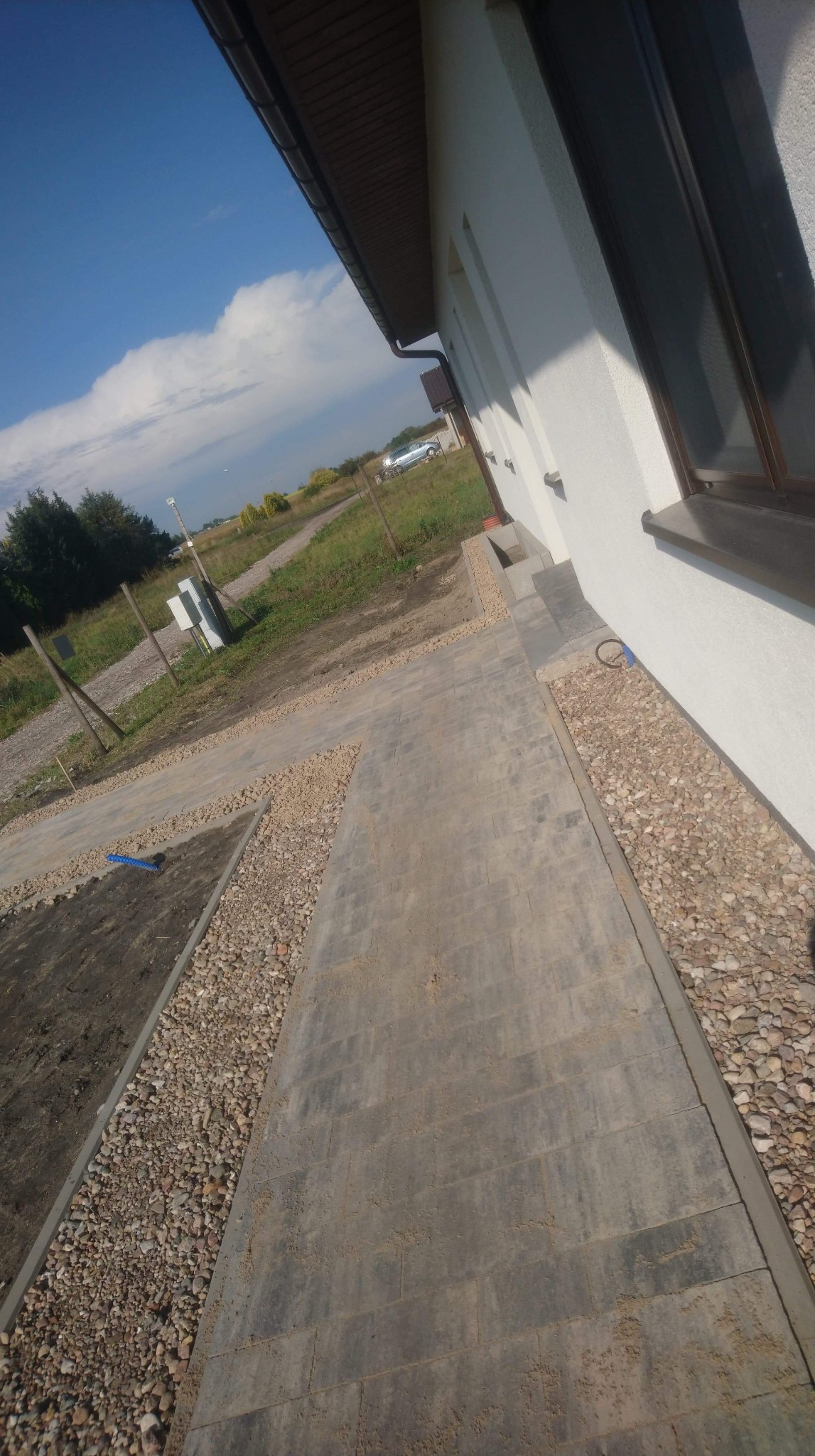 Świeżo ułożona szara kostka brukowa przy białej ścianie budynku, oddzielona od trawnika pasem drobnych kamieni, widoczny fragment nieukończonej powierzchni z przygotowanym podłożem.