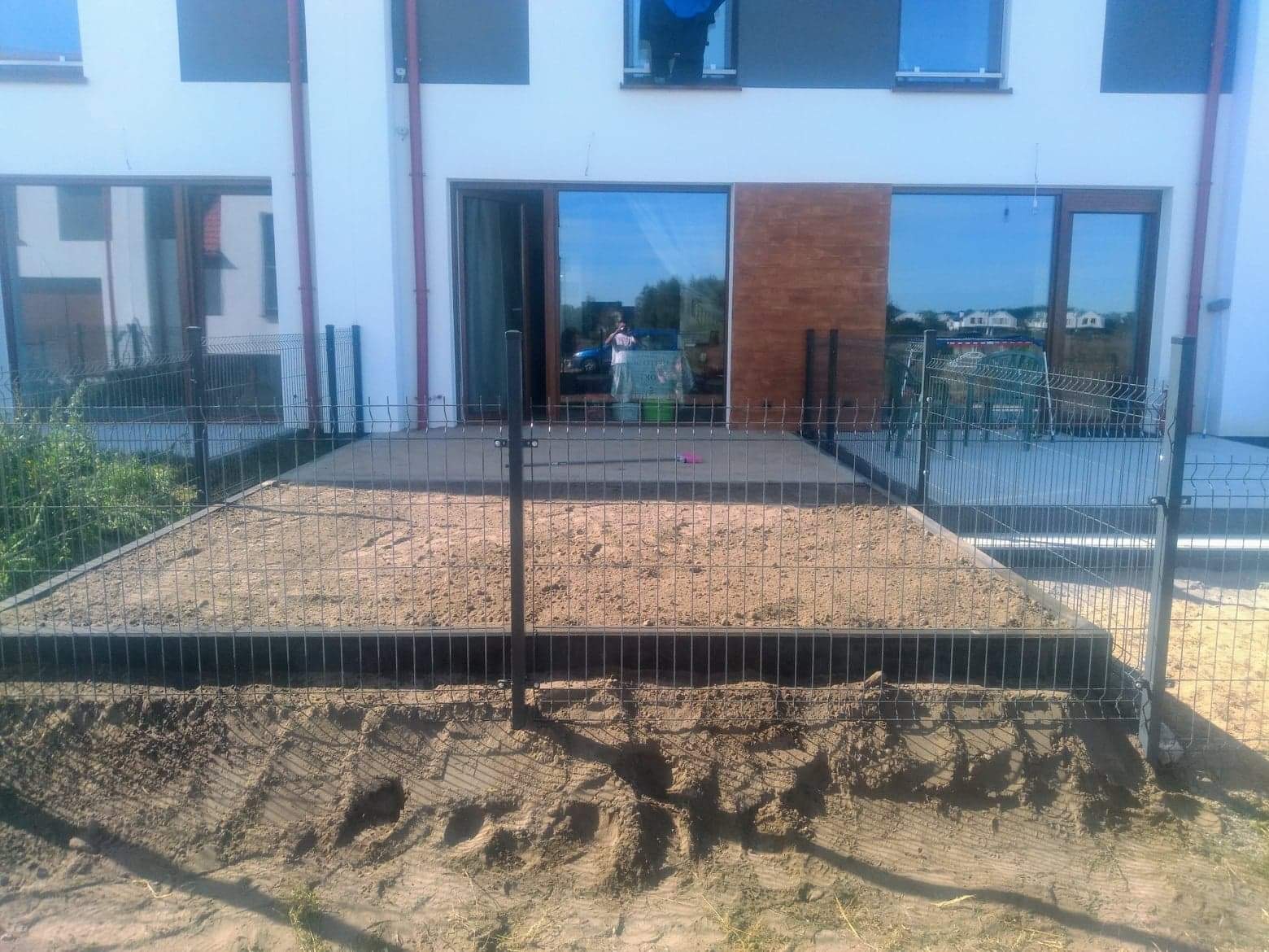 Przygotowanie podłoża pod taras: wyrównany teren z piaskiem i obrzeżami, ogrodzony siatką, widok na nowoczesny dom z dużymi oknami.