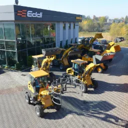 Plac z nowymi, żółtymi maszynami budowlanymi, w tym wózkami widłowymi, na tle budynku firmy Eddi z dużymi oknami wystawowymi i logo.