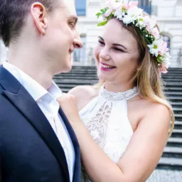 Uśmiechnięta kobieta w białej sukience i kwiecistym wianku poprawia krawat mężczyźnie w czarnym garniturze na tle schodów i budynku.